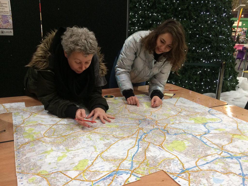 Two women lean over a map on a table, one has short, curly grey hair and a pen in her hand, the other has long brown hair and is pointing and smiling 